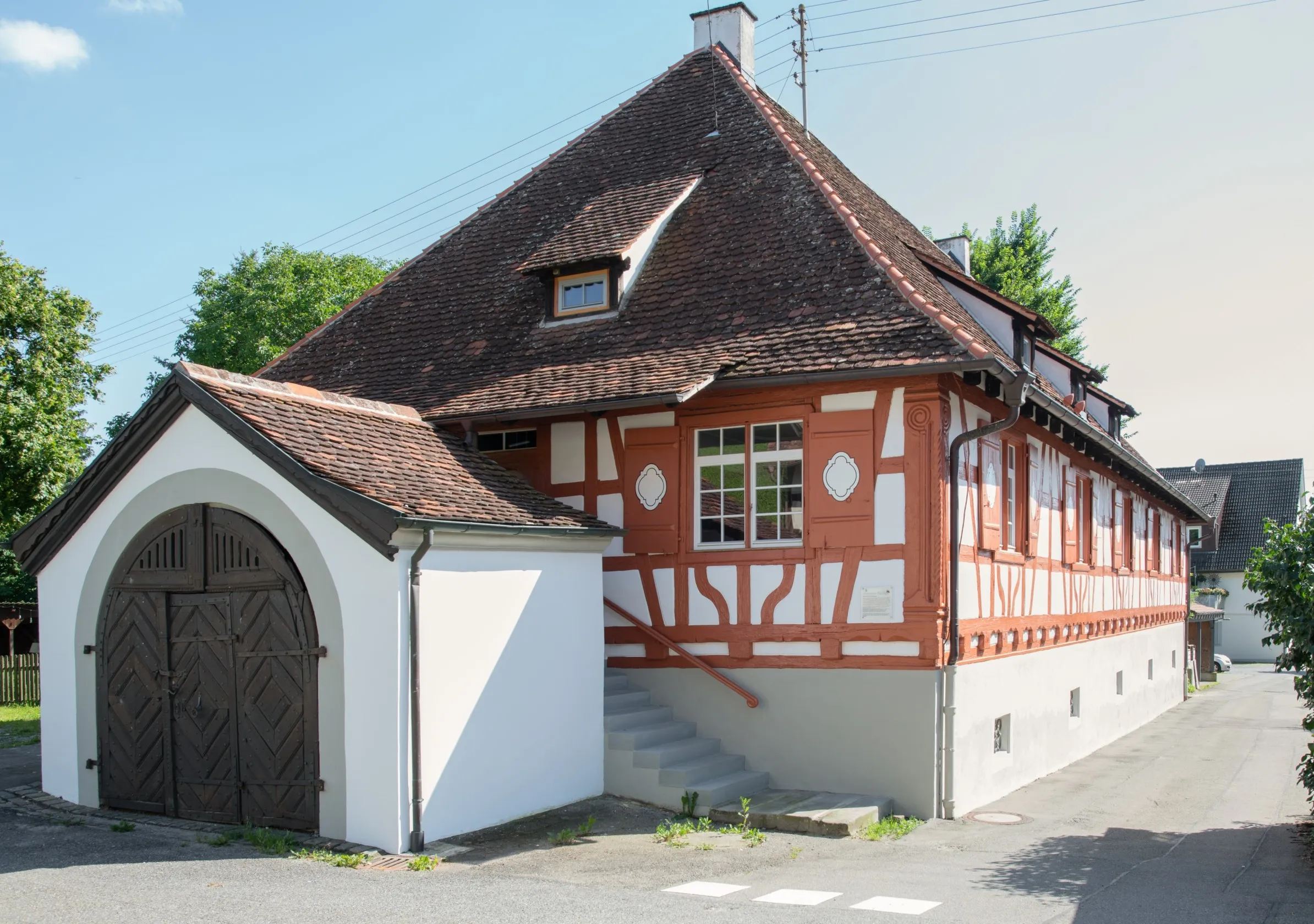 Außenansicht des Jägerhaus in Bermatingen. Altbau mit rotem Fachwerk, weißen Gefachen, Dachziegel aus Biberschwanzziegeln und dem Abgang in das Kellergewölbe im Vordergrund, Bäume im Hintergrund