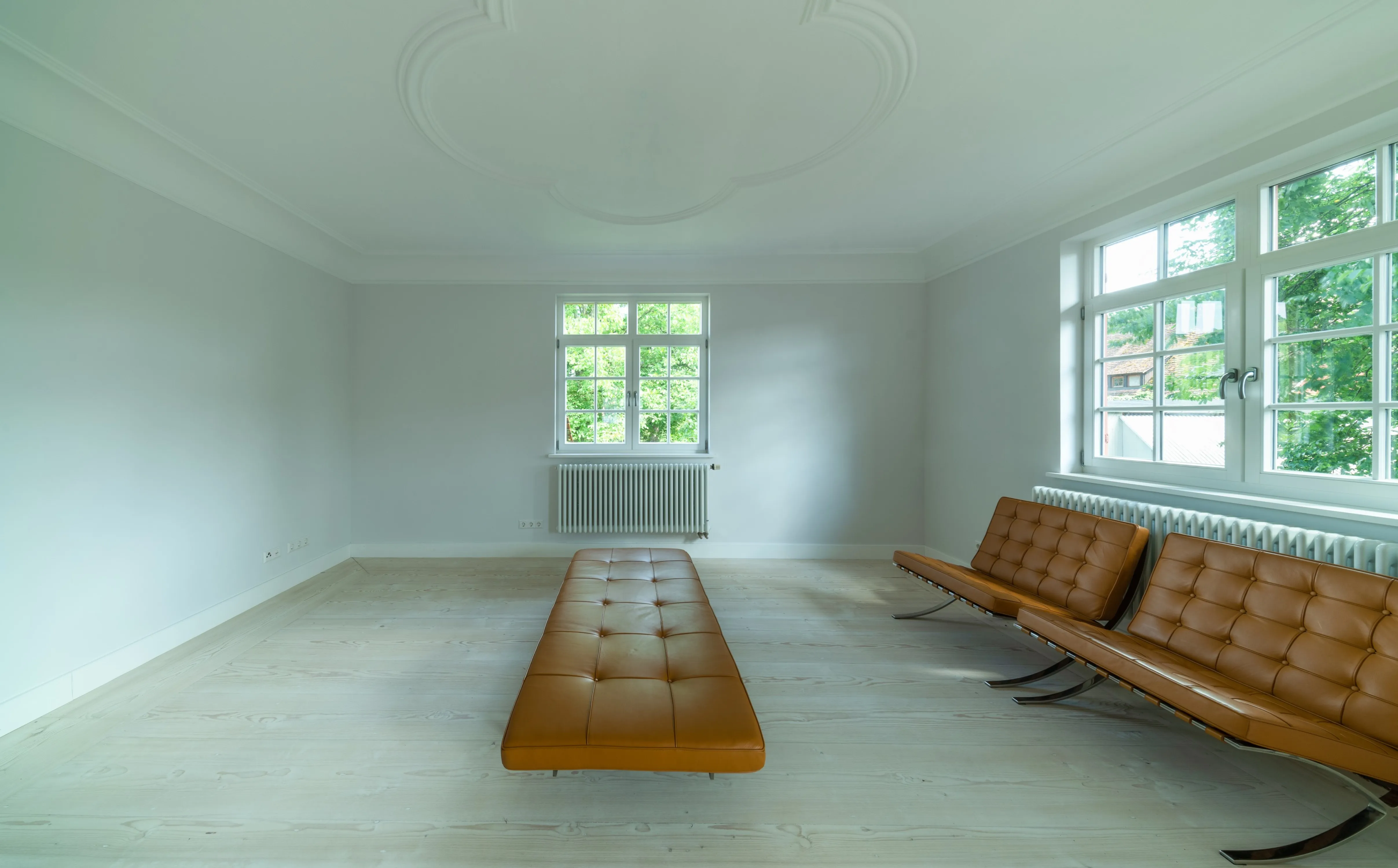 Antmosphärischer Innenraum mit Stuckdecken und zwei Fenstern. Im Raum stehen zwei orangefarbene / braune Barcelona Chairs und Ottomane in Raummitte