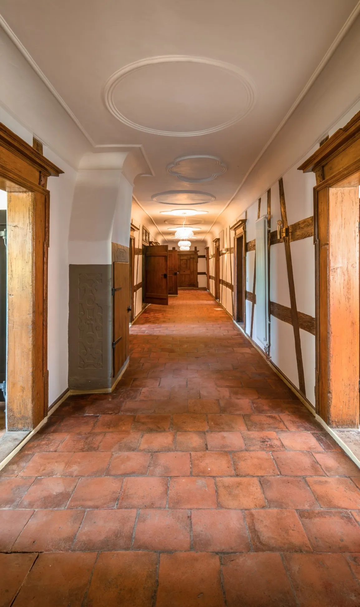 Langer Flur mit roten Tonfliesen, Fachwerkwänden und Stuckdecke, Türen rechts und links zu den Zimmern