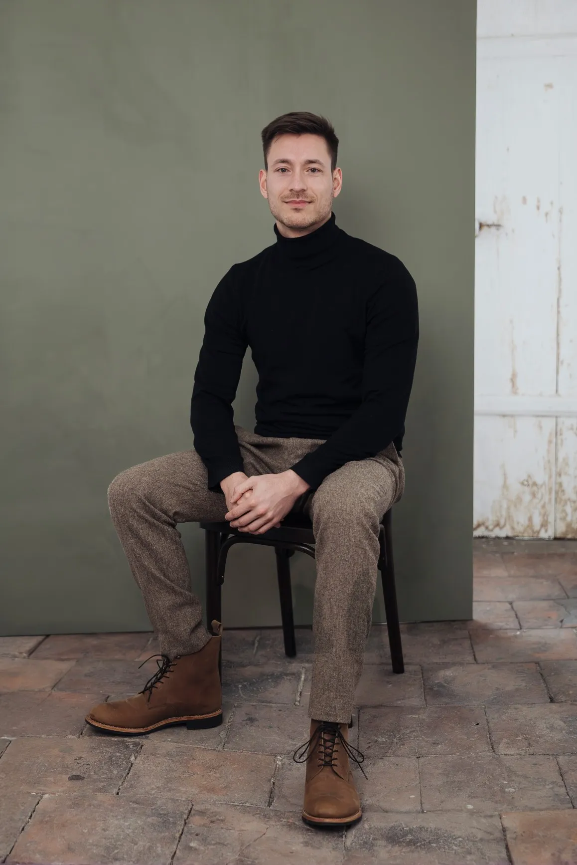 Eric Lenz Architekt und Sachverständiger für Immobilienbewertung trägt einen schwarzen Rollkragenpullover, braune Hose und Schuhe und sitzt auf einem alten Thonet Stuhl vor einer grünen Leinwand in einem Altbau auf der Insel Lindau